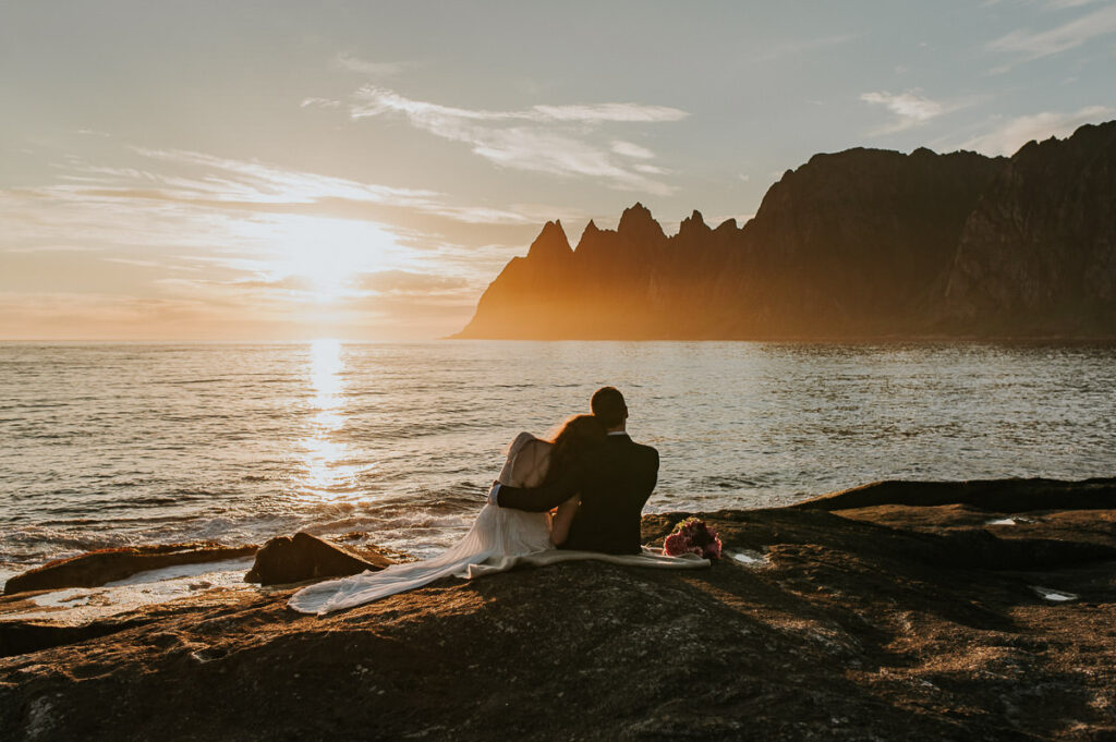 Bride and groom sitting on the rocks and enjoying midnight sun and gorgeous mountain views of Senja island in Northern Norway