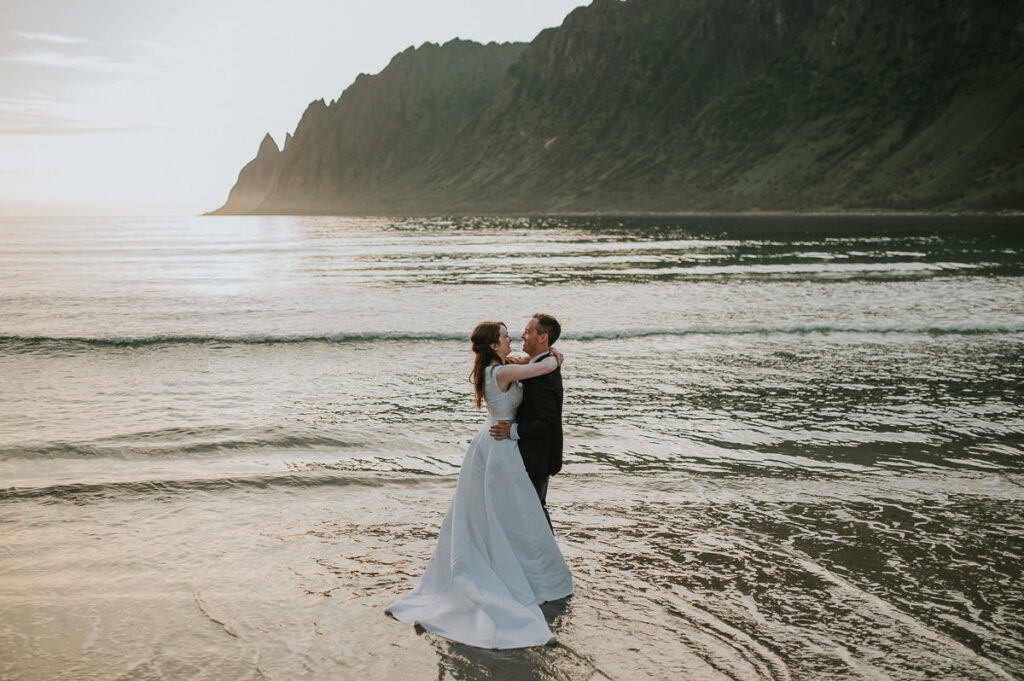 Bride and groom dipping their toes in the arctic water on a beach on Senja island in Norway. They're wearing wedding attire and getting wet