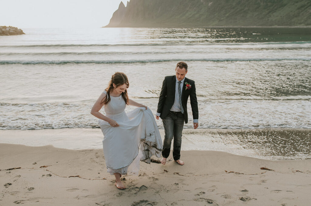 Bride and groom dipping their toes in the arctic water on a beach on Senja island in Norway. They're wearing wedding attire and getting wet