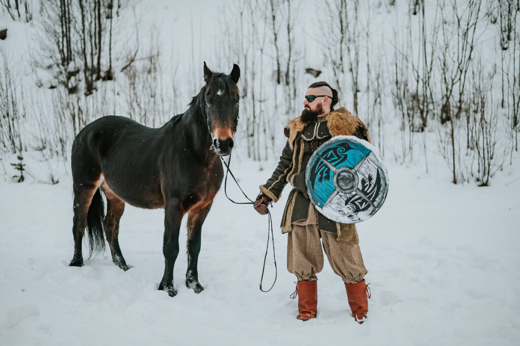 Male viking ina  cool outfit pets a horse in front of a snowy winter landscape in Norway