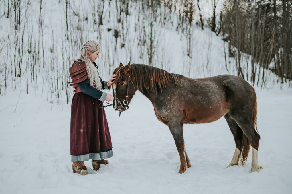 Viking shieldmaiden wearing authentic viking outfit with apron dress and leather armor pets a horse in front of a snowy winter landscape in Norway