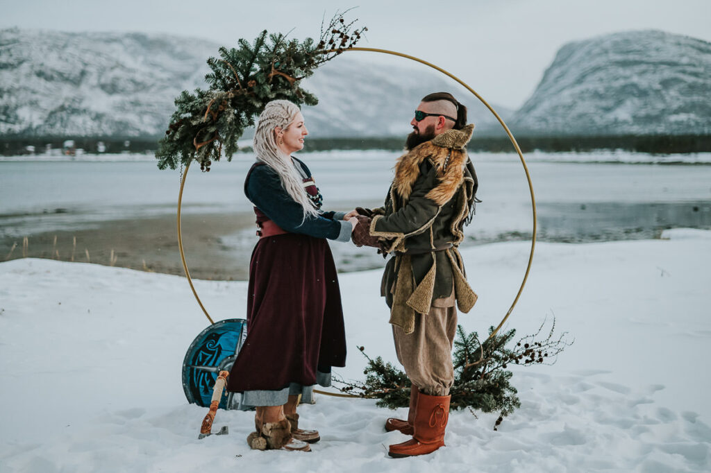 Viking style wedding ceremony in Alta Norway - beautiful couple in cool viking outfits are getting married under a round floral arch decorated with antlers and winter decor. Behind them is a beautiful snowy landscape with mountains and sea