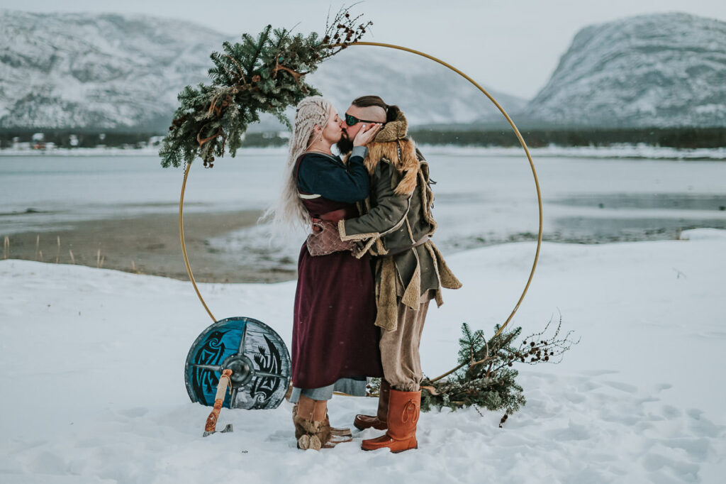 Viking style wedding ceremony in Alta Norway - beautiful couple in cool viking outfits are getting married under a round circular floral arch decorated with antlers and winter decor. Behind them is a beautiful snowy landscape with mountains and sea