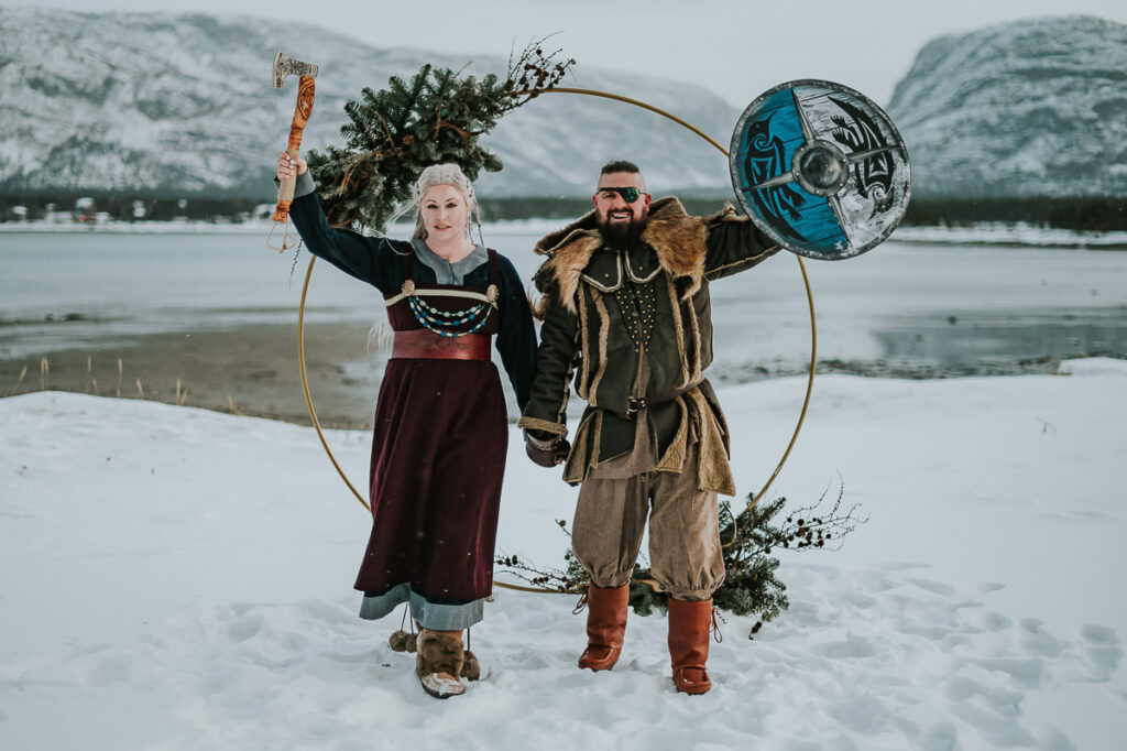 Viking style wedding ceremony in Alta Norway - beautiful couple in cool viking outfits are getting married under a floral arch decorated with antlers and winter decor. Behind them is a beautiful snowy landscape with mountains and sea