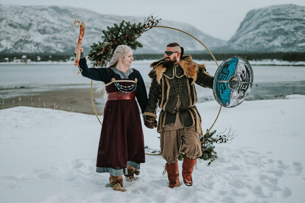 Viking style elopement ceremony in Alta Norway - beautiful couple in cool viking outfits are getting married under a floral arch decorated with antlers and winter decor. Behind them is a beautiful snowy landscape with mountains and sea