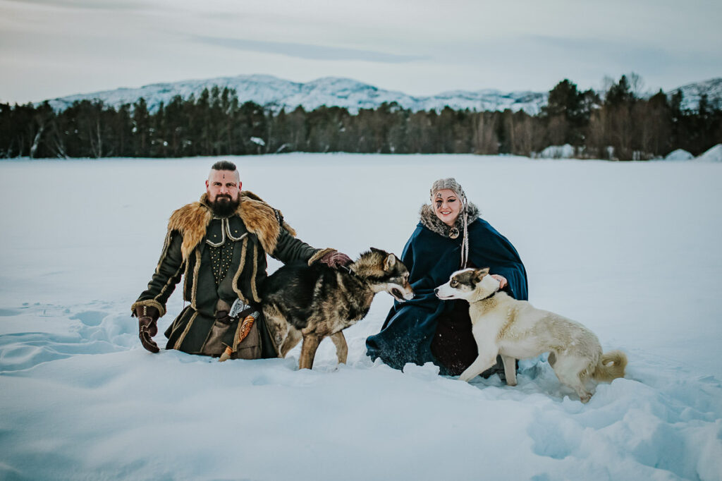 Viking couple cuddling with husky dogs in front of a snowy winter landscape