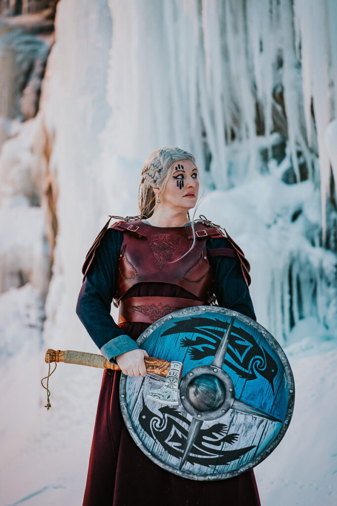 Viking shieldmaiden portrait in front of frozen waterfalls in Norway. She is pozing with an axe and shield in her hands and is wearing a leather armor 