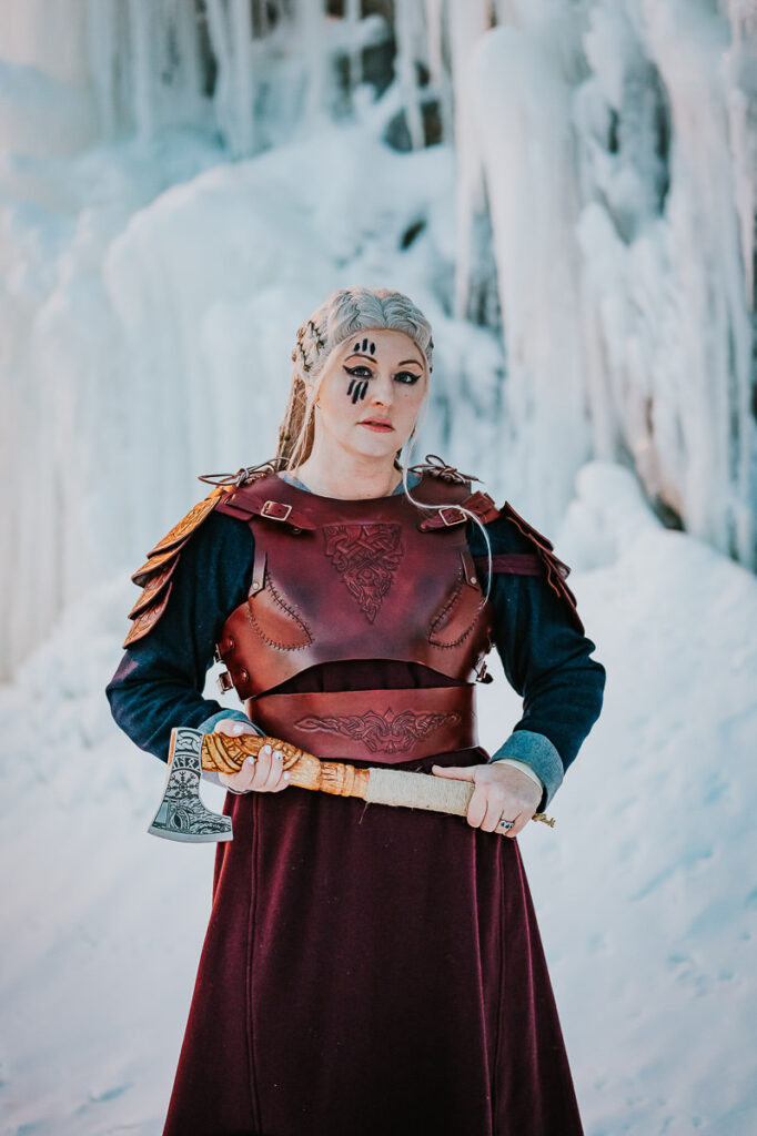 Viking shieldmaiden portrait in front of frozen waterfalls in Norway. She is pozing with an axe and is wearing a leather armor 