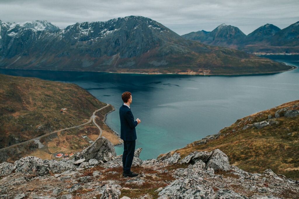 A groom in a navy blue wool suit waiting for his bride on a mountaintop in Tromsø overlooking a great fjord view