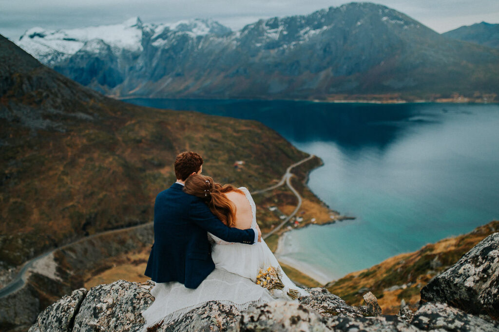 Bride and groom sitting on a rock and hugging each other while admiring a great view with snowy-capped mountains and the sea in the background - hiking elopement in Tromsø