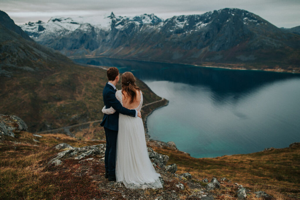 Bride and groom hugging each other on a mountaintop while admiring a great view with snow-capped mountains and the sea in the background - hiking elopement in Tromsø