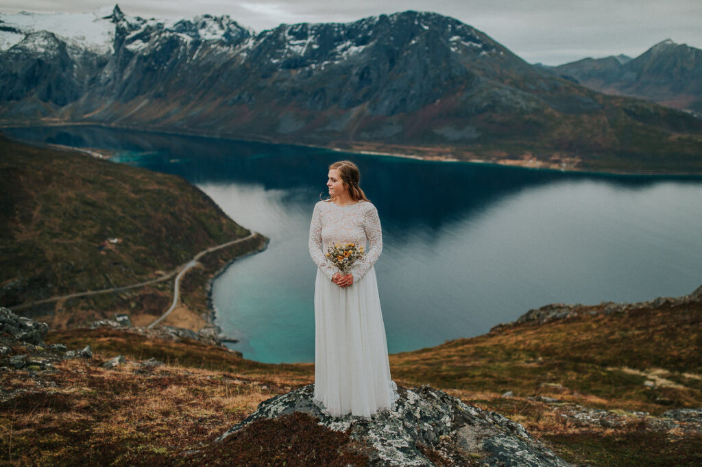 Gorgeous bride in a lace wedding gown holding a yellow dried flowes bouquet on a mountaintop in Norway with a great fjord view