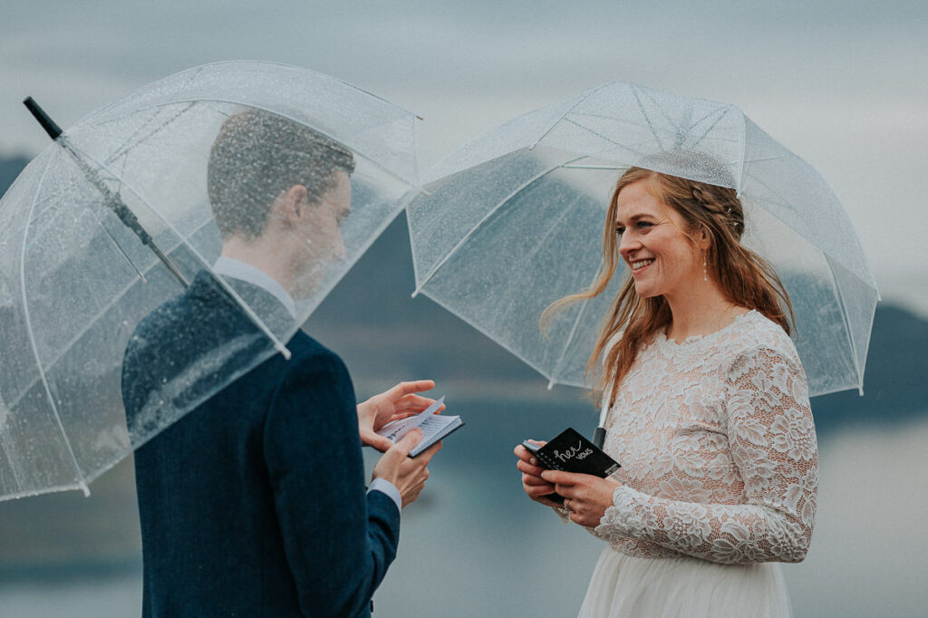 Rainy day fall elopement in Tromsø. Bride and groom reading their vows to each other on a mountaintop while hiding under clear umbrellas