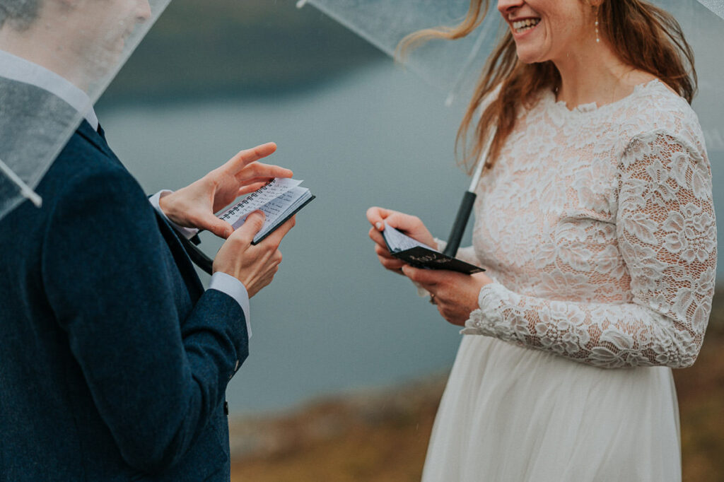 Rainy day fall elopement in Norway. Bride and groom reading their vows to each other on a mountaintop while hiding under clear umbrellas