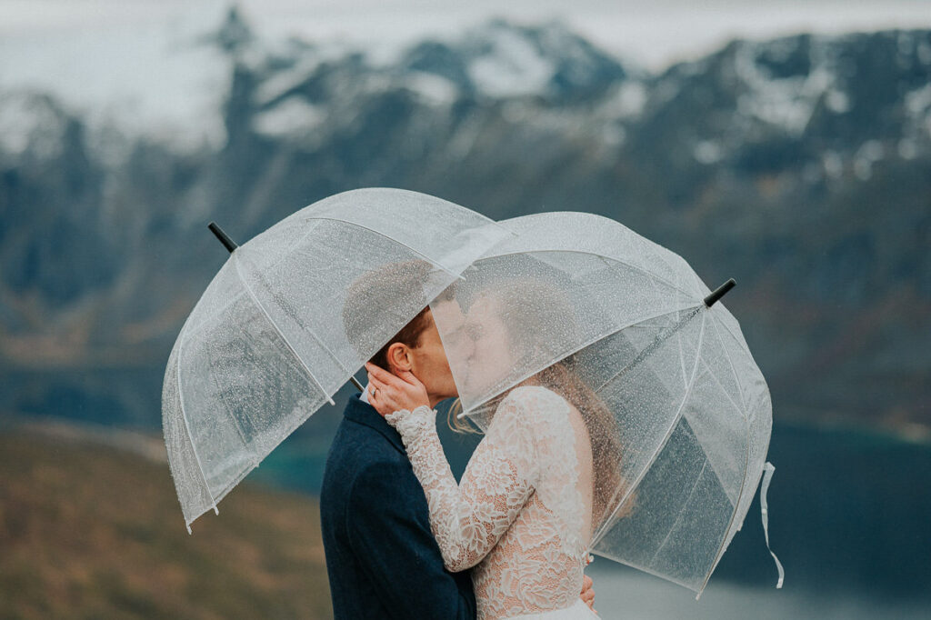 Rainy day fall elopement in Tromsø. Bride and groom kissing each other on a mountaintop while hiding under clear umbrellas
