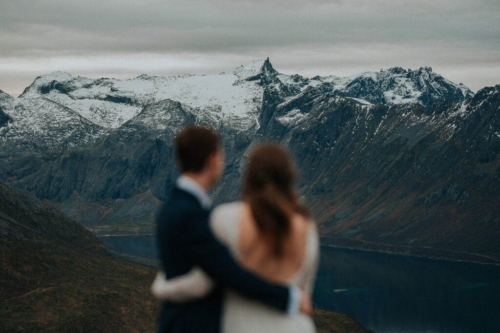 Bride and groom hugging each other on a mountaintop while admiring a great view with snow-capped mountains and the sea in the background