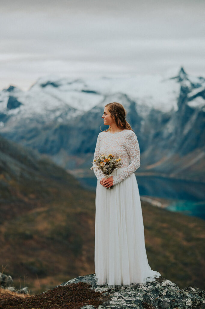 Gorgeous bride in a lace wedding gown holding a yellow dried flowes bouquet on a mountaintop in Tromsø with a great fjord view