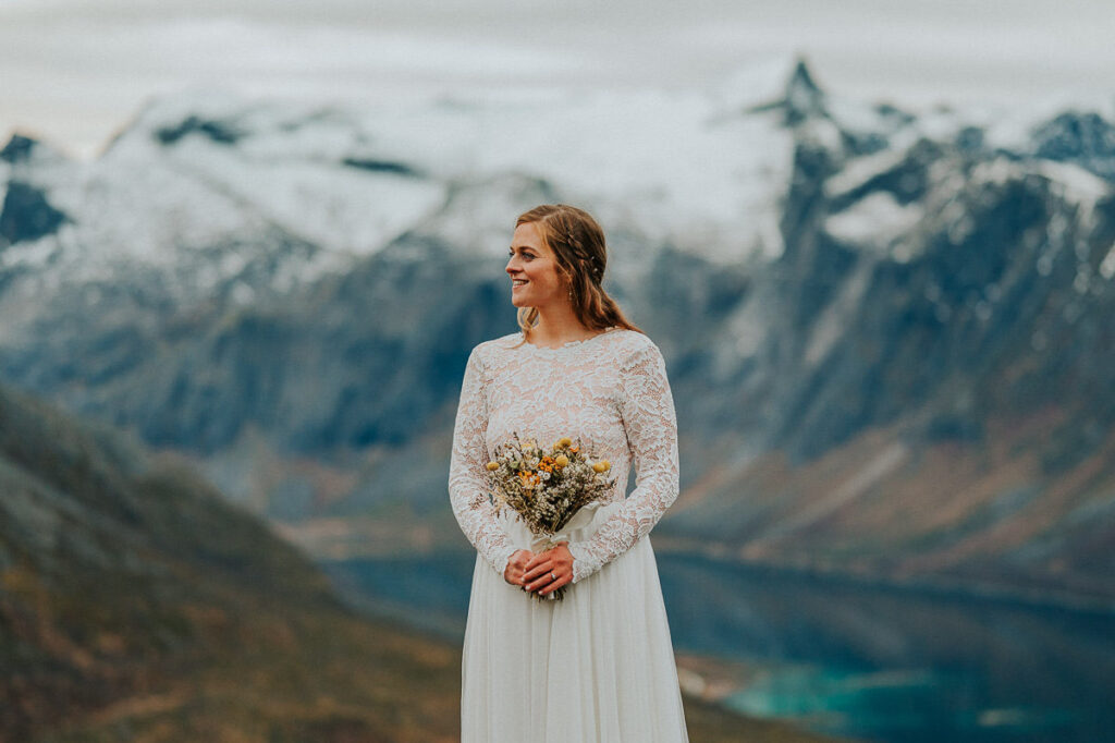Gorgeous bride in a lace wedding gown standing on a mountaintop in Tromsø with a great fjord view