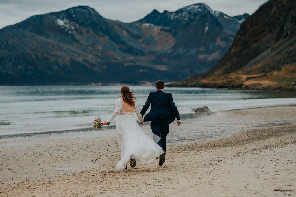 Bride and groom holding hands and running on the beach towards the sea on the day of their hiking elopement in Tromsø Norway