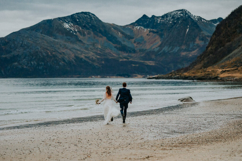 Bride and groom holding hands and running on the beach towards the sea on the day of their hiking elopement in Tromsø Norway