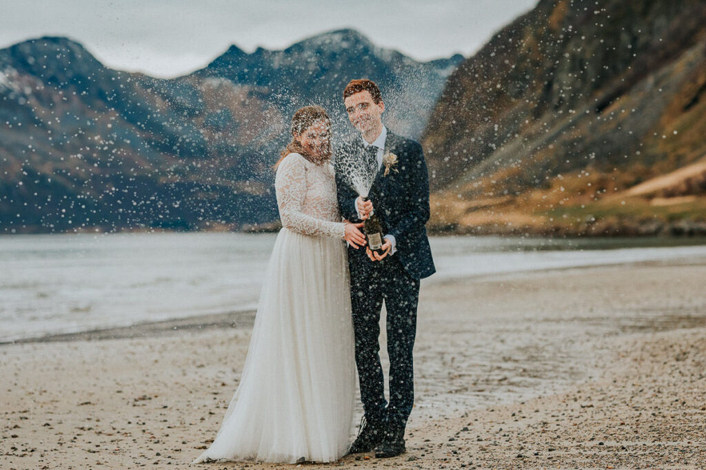Bride and groom popping up and spraying champagne on a beach with a mountainview  on the day of their hiking elopement in Tromsø Norway