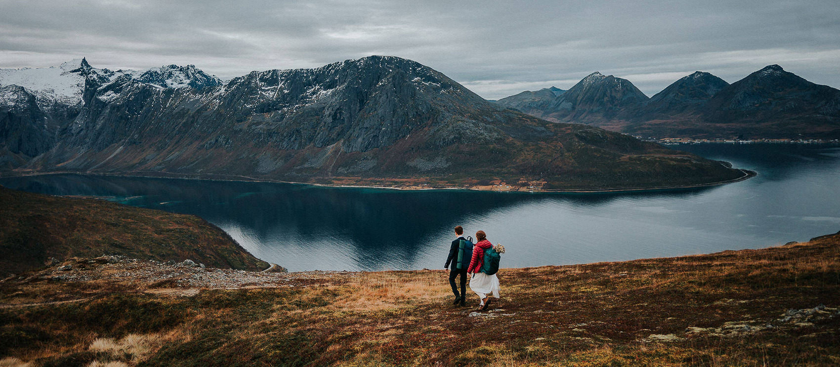 Bride and groom hiking in the mountains of Tromsø on their elopement day. There is a view of snow-capped mountains in the background and the fall colors on the ground