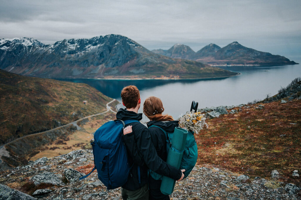 Bride and groom hiking to a mountaintop on the day of their elopement in Tromsø. They're wearing hiking clothes and carry flowes and umbrellas in their backpacks