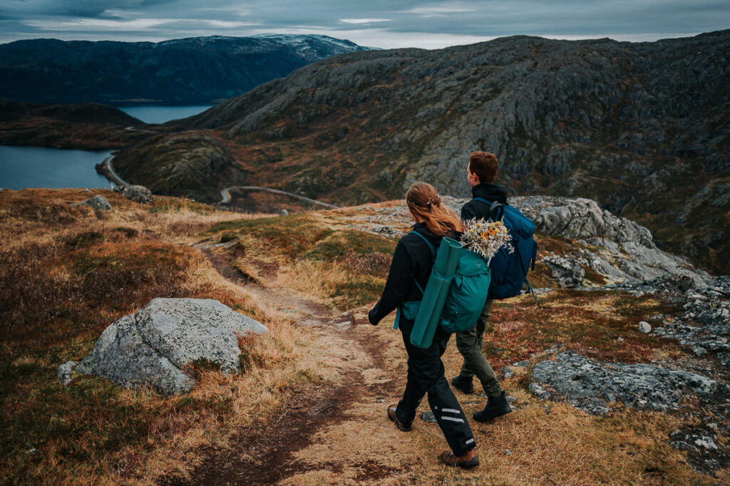 Bride and groom hiking to a mountaintop on the day of their elopement in Tromsø. They're wearing hiking clothes and carry flowes and umbrellas in their backpacks