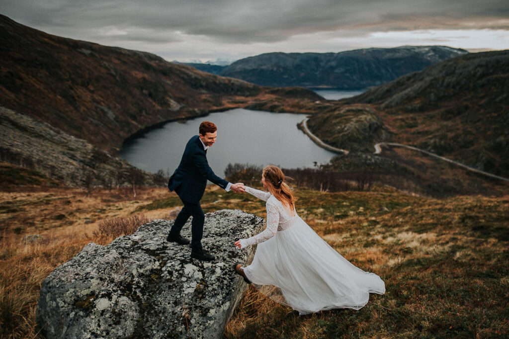Epic bride jumping on a big rock in front of a beautiful fall mountain landscape in Tromsø and the groom is giving her a hand - mountain hiking elopement in Tromsø
