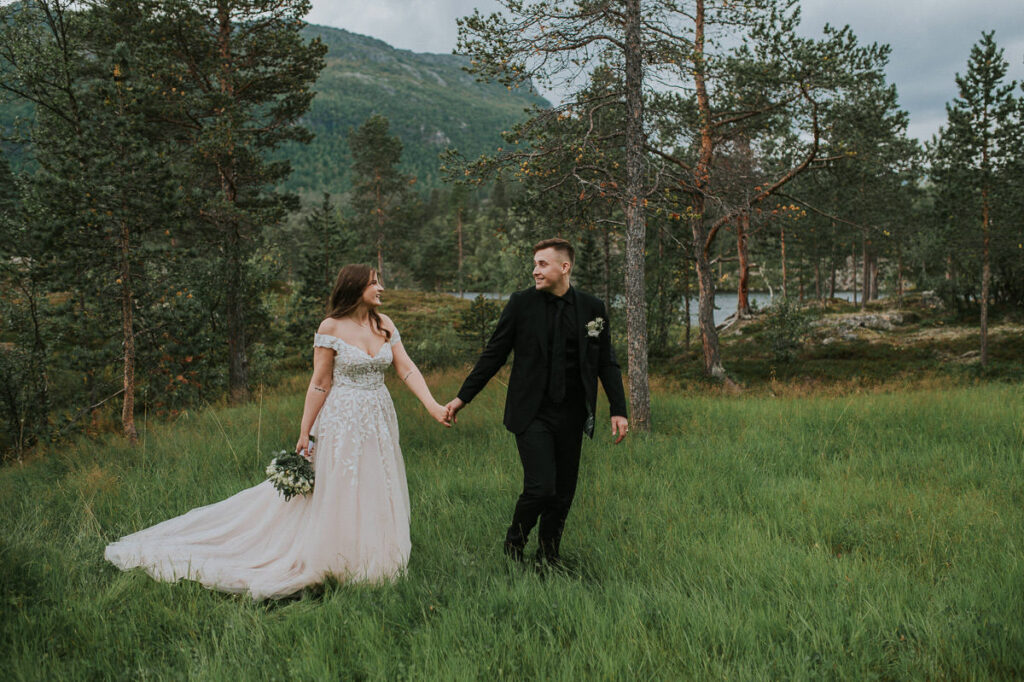Brudepar holder hverandre i hendene mens de spaserer på grønt gress på en lund i skogen
