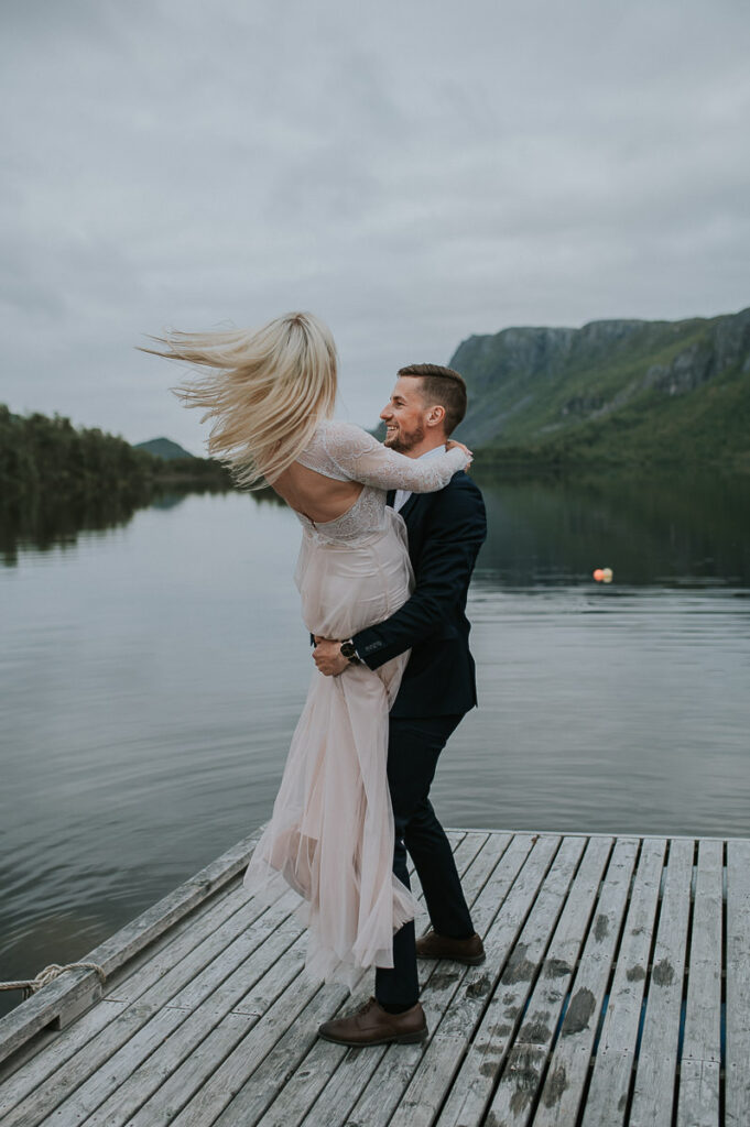 Brudepar står på brygga foran et innsjø og fjell i Alta, Finnmark. Brudgommen løftet bruden sin og snurrer henne rundt mens hun flirer