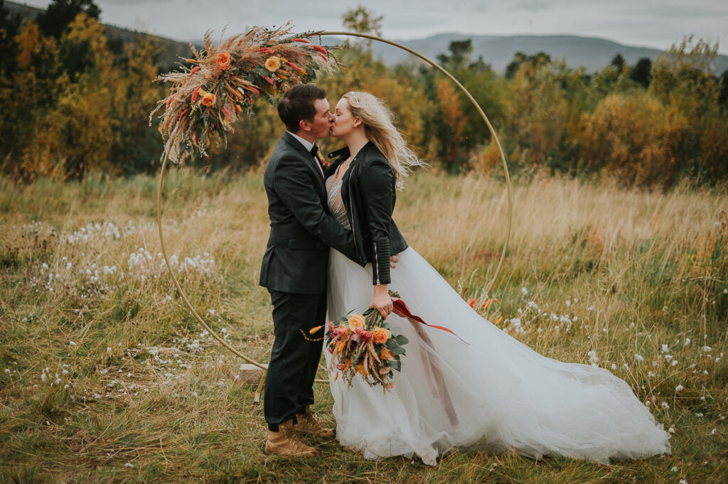 Vakkert brudepar kysser i et stråfelt i Alta under en rund blomsterbua med tørket blomster - beste foto locations for et høstbryllup i Alta