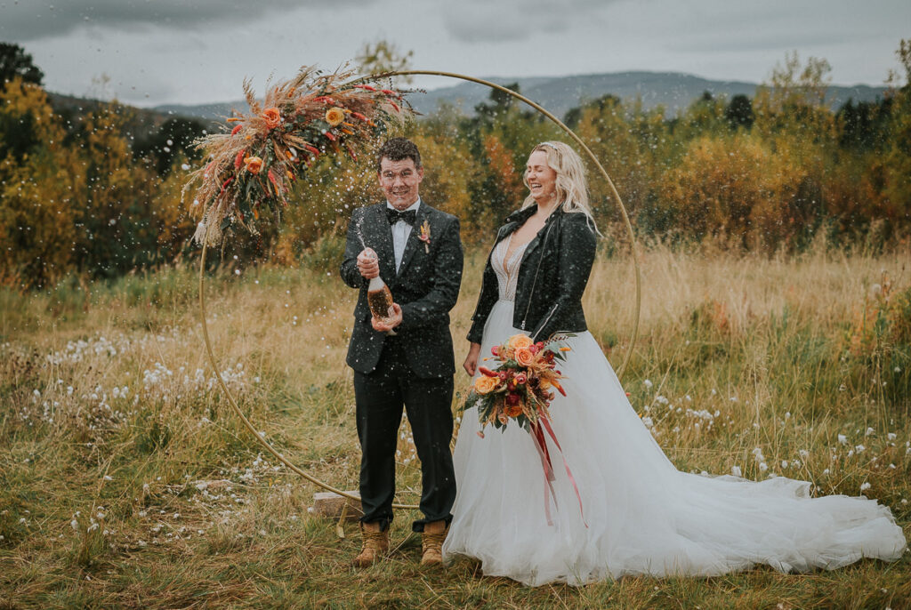 Nydelig brudepar popper champagne foran et vakkert utsikt til fjell og gyldent stråfelt med en rund blomsterbua i bakgrunn