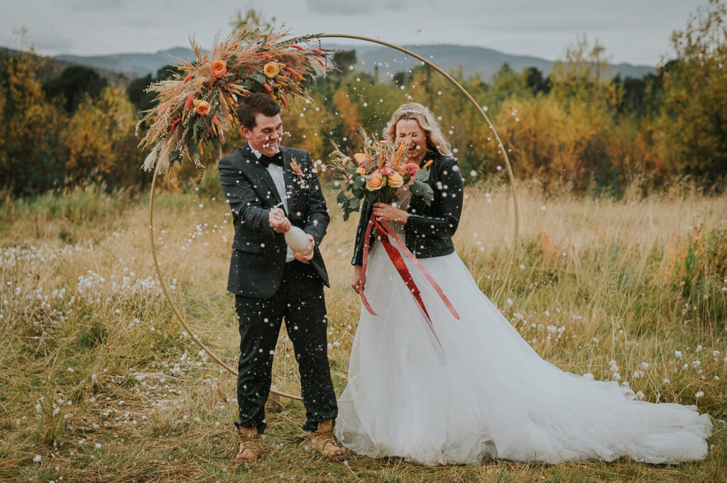Nydelig brudepar popper champagne foran et vakkert utsikt til fjell og gyldent stråfelt med en rund blomsterbua i bakgrunn