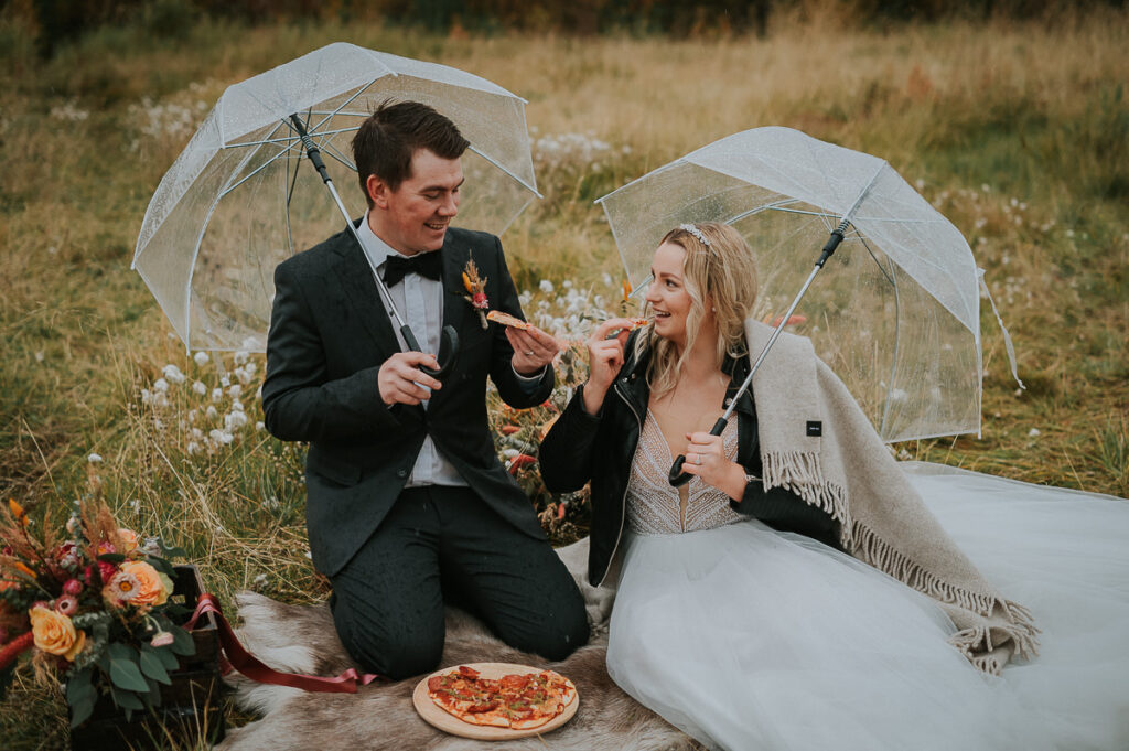 Brudepar sitter utendørs under paraply og spiser pizza på bryllupsdagen sin