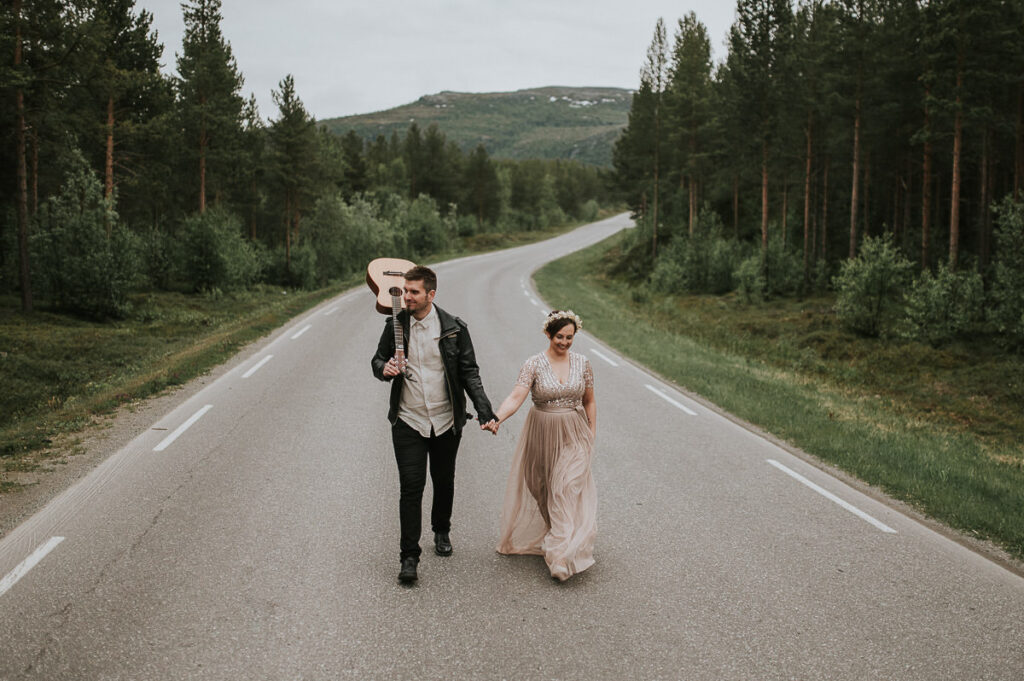 Nydelig brudepar spaserer midt på veien i Alta Norge. Brudgommen holder gitar på skuldre og har på seg skinnjakke 