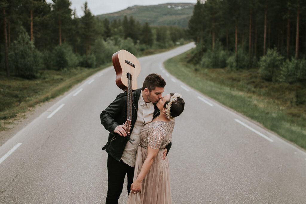Nydelig brudepar spaserer midt på veien i Alta Norge. Brudgommen holder gitar på skuldre og har på seg skinnjakke 