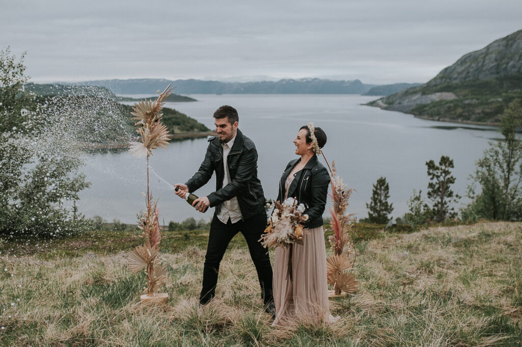 Nydelig brudepar popper champagne foran et vakkert utsikt til fjord og fjell med en  blomsterbua dekorert med tørkede blomster i bakgrunn