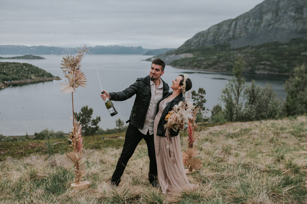 Nydelig brudepar popper champagne foran et vakkert utsikt til fjord og fjell med en  blomsterbua dekorert med tørkede blomster i bakgrunn
