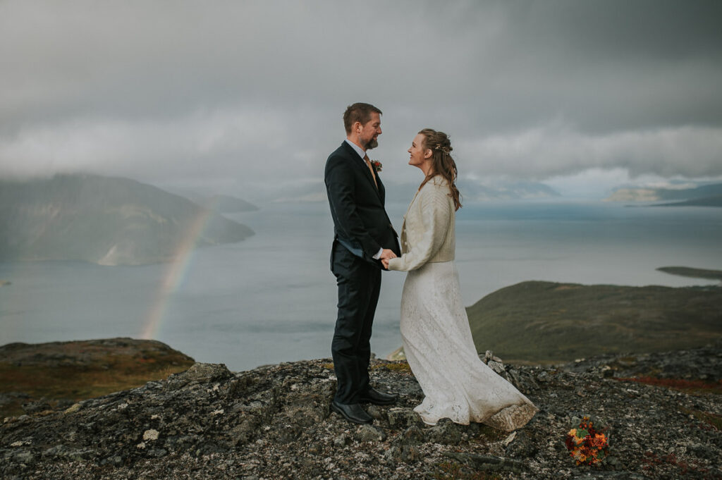 Brudepar holder hverandres hender med en nydelig regnbue og fjell utsikt i bakgrunn