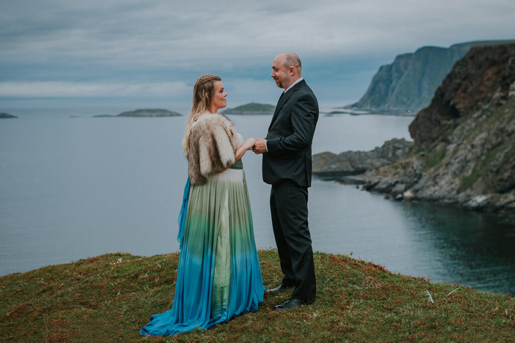 Bride and groom are walking among beautiful landscapes on Sørøya island in Northern Norway on the day of their midnight sun elopement. The bride is wearing a beautiful ivory green blue dress with ombre effect
