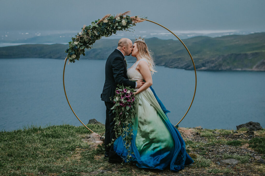 Bride and groom kissing on their  midnight sun elopement ceremony on Sørøya island in Norway. The couple is standing in front of a round wedding arch decorated with blue and green flowers. Bride is wearing a blue green wedding dress and has a viking inspired hair style