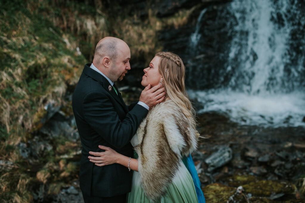 Bride and groom posing in front of a waterfall in Sørøya island in Norway. Bride is wearing blue green wedding gown and has a viking inspired hair style