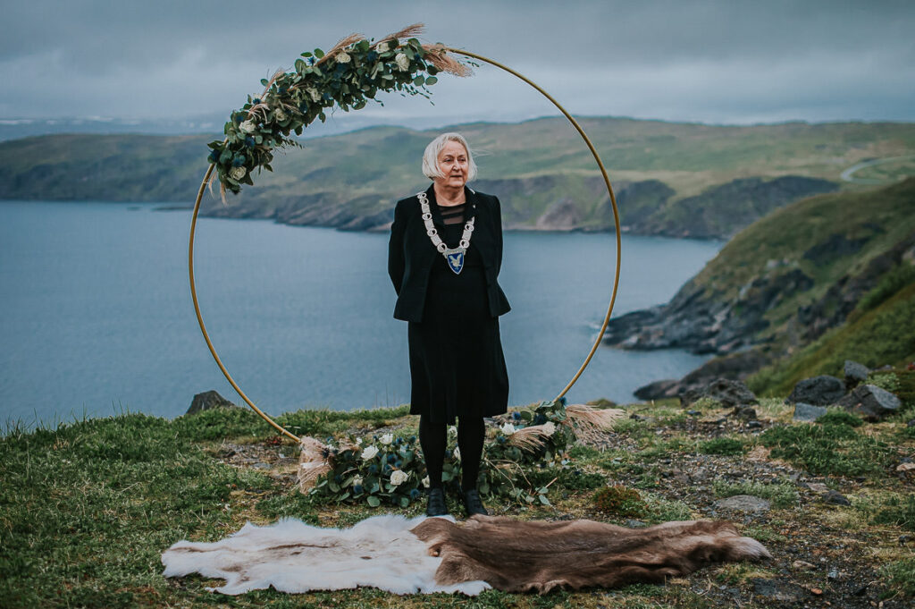 Mayor of Hasvik in Sørøya is waiting for the bride and groom in front of a round floral arch standing in front of a beautiful nordic landscape in Norway. The arch is decorated with blue and green flowers 