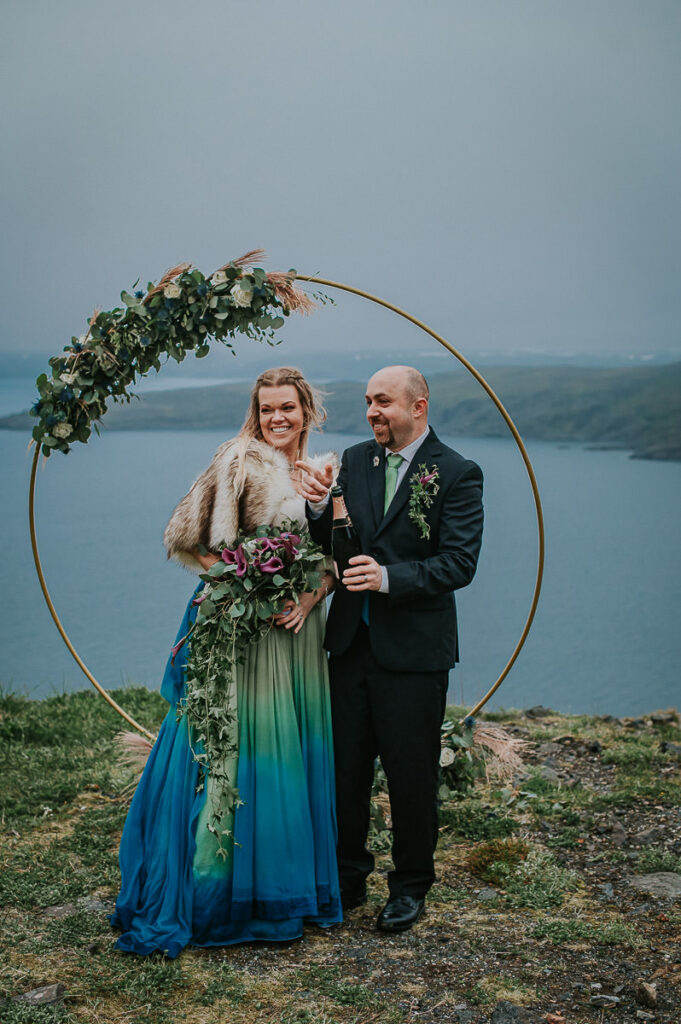 Bride and groom popping and drinking champagne after their elopement ceremony on Sørøya island in Norway. The couple is standing in front of a round wedding arch decorated with blue and green flowers. Bride is wearing a blue green wedding dress and has a viking inspired hair style