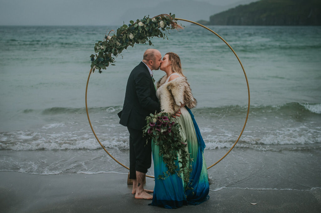 Bride and groom kissing in front of a round floral arch decorated  with blue and green flowers in the sea on a foggy night . They are dipping toes in the cold water and enjoying their midnight sun elopement in Sørøya island in Norway