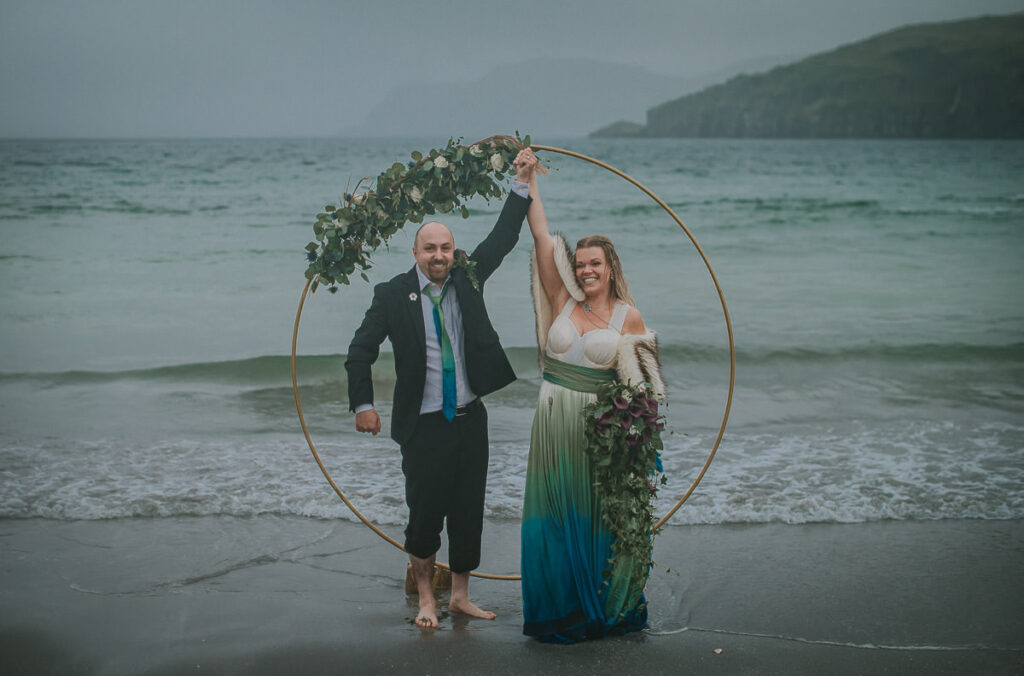 Bride and groom popping champagne in front of a round floral arch decorated  with blue and green flowers in the sea on a foggy night . Their feet are dipped in the cold water and they are enjoying their midnight sun elopement in Sørøya island in Norway