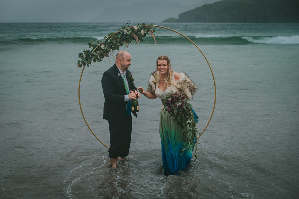 Bride and groom popping champagne in front of a round floral arch decorated  with blue and green flowers in the sea on a foggy night . Their feet are dipped in the cold water and they are enjoying their midnight sun elopement in Sørøya island in Norway