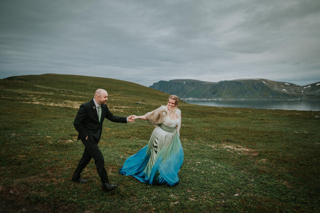 Bride and groom are walking among beautiful landscapes on Sørøya island in Northern Norway on the day of their midnight sun elopement. The bride is wearing a beautiful ivory green blue dress with ombre effect