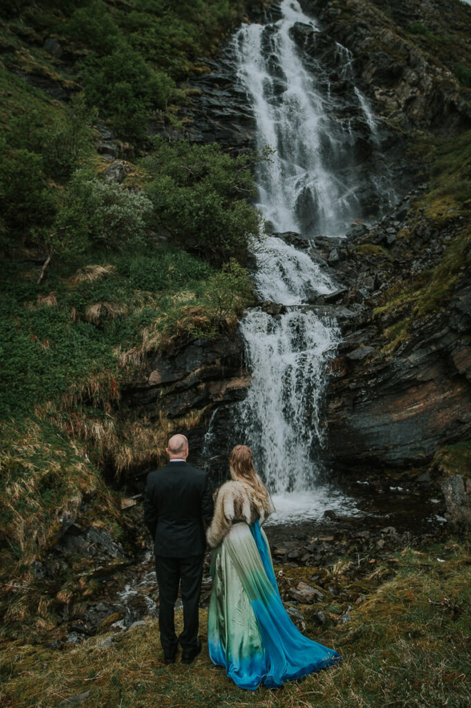 Bride and groom posing in front of a waterfall in Sørøya island in Norway. Bride is wearing blue green wedding gown and has a viking inspired hair style